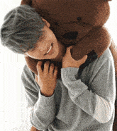 a young man in a grey sweater is hugging a large teddy bear