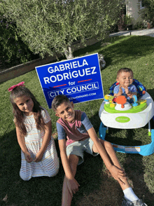 three children sit in front of a gabriela rodriguez sign for city council