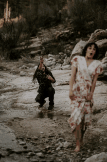 a woman in a floral dress is standing in a stream while a man kneeling in the water looks on