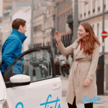 a woman in a trench coat waves to a man in a blue jacket in front of a white car that says swafiets