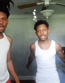 two men in white tank tops are standing next to each other in a room with a ceiling fan