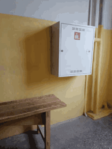 a wooden bench sits in front of a yellow wall and a white box