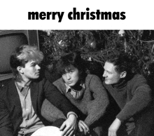 three men are sitting in front of a christmas tree with merry christmas written above them