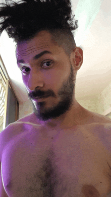 a shirtless man with a beard and a bun on his head looks at the camera