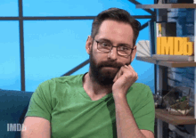 a man wearing glasses and a green shirt is sitting in front of a shelf with a sign that says imdb