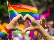 a couple of hands making a heart shape with a rainbow flag in the background