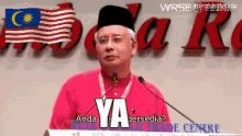 a man stands at a podium with a flag in the background and says ya anda bersedia