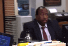 a man in a suit and tie is sitting at a desk in an office with a water cooler in the background .