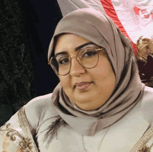 a woman wearing glasses and a hijab stands in front of a red white and blue flag that says bloom