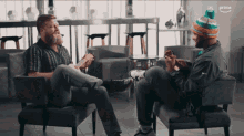 two men are sitting in chairs and one is playing a guitar while the other is talking