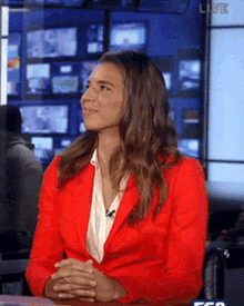 a woman in a red jacket is sitting at a table with her hands folded in front of a live screen