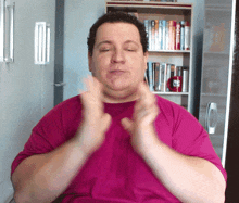 a man wearing a pink shirt is clapping his hands in front of a bookshelf with a red mug that says coca cola on it