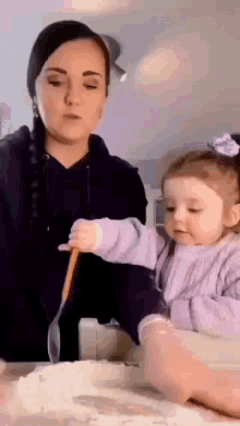 a woman and a little girl are playing with flour on a table . the little girl is holding a wooden spoon .