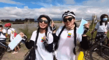 two women wearing helmets and sunglasses are standing next to each other on a dirt road with bicycles in the background .