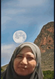 a woman wearing a hijab stands in front of a mountain and a full moon