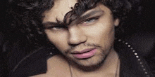 a man with curly hair and a beard is wearing a black jacket and necklace .