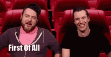 two men are sitting in red seats in a theater and one of them is smiling .