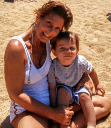 a woman in a white tank top sits on the beach holding a child