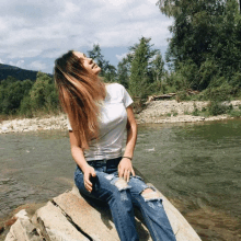 a woman in a white shirt and ripped jeans sits on a rock by a river