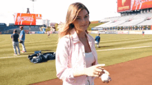 a woman in a pink jacket is holding a baseball on a baseball field with an ad for coca cola behind her