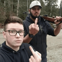 two men are standing next to each other and one of them is holding a rifle and giving the middle finger .