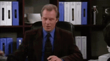 a man in a suit and tie is sitting at a desk in front of a shelf filled with binders .