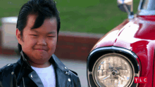 a young boy in a leather jacket stands in front of a red car with tlc written on the bottom