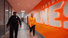 a police officer and a woman walk down a hallway in front of a wall that says " it 's basket ball "