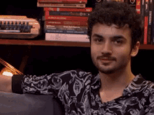 a man with curly hair and a beard is sitting in front of a bookshelf with books on it .