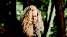 a woman with blonde curly hair stands in a forest