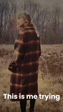 a woman in a plaid coat is standing in a field with the words " this is me trying " above her