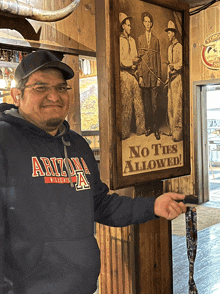 a man wearing an arizona sweatshirt stands in front of a no ties allowed poster