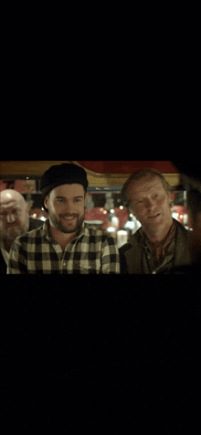 a man wearing a plaid shirt and a black hat smiles while standing next to two other men