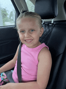 a little girl wearing a pink tank top and a seat belt smiles for the camera