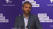 a man in a suit stands in front of a purple sign that says " staying apart keeps us together "