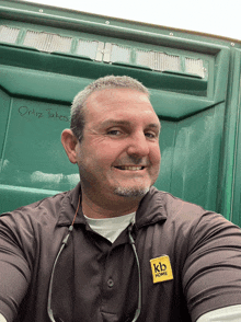 a man wearing a kb home shirt smiles in front of a green stall