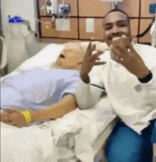 a man is sitting next to a man in a hospital bed giving the middle finger .