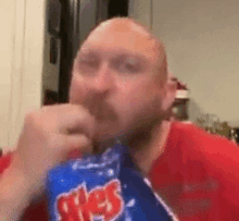 a bald man in a red shirt is eating a bag of chips .