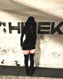 a woman in a black dress stands in front of a wall that has the word hiek painted on it