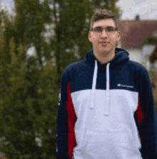 a young man wearing a hoodie and glasses is standing in front of a tree .