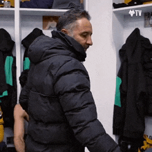 a man in a black jacket is standing in a locker room with a tv logo on the wall above him