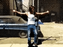 a man in a white tank top and jeans is standing in front of a chain link fence with his arms outstretched