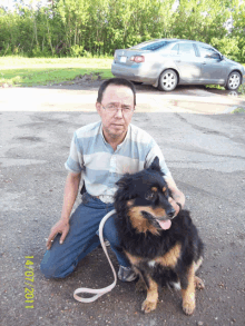 a man kneeling down next to a dog with a car parked in the background