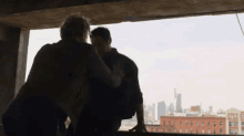 two men are standing next to each other in front of a window with a city skyline in the background .