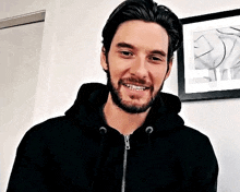 a man with a beard is wearing a black hoodie and smiling in front of a picture on a wall .