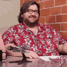 a man wearing a red shirt with flowers on it sits at a desk in front of a brick wall