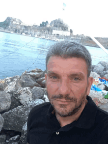 a man in a black shirt stands in front of a body of water and rocks