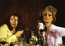 two women are sitting at a table holding wine bottles and glasses