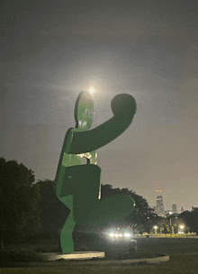 a large green sculpture in a park with a city skyline in the background