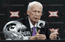 a man in a suit and tie is speaking into a microphone in front of a big 12 conference sign